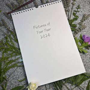 Englisches Cover „Pictures of Your Year 2026“ in Handschrift-Optik – ringgebundener A3-Blankokalender mit Wandaufhängung, umgeben von Blumen und Gräsern