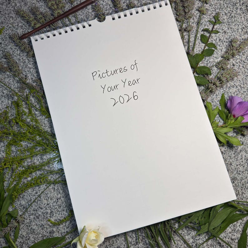 Englisches Cover „Pictures of Your Year 2026“ in Handschrift-Optik – ringgebundener A3-Blankokalender mit Wandaufhängung, umgeben von Blumen und Gräsern