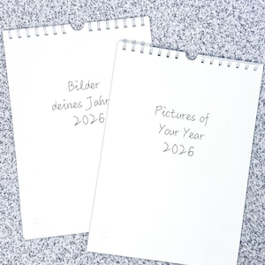 Op de afbeelding: Twee blanco witte kalenders met spiraalbinding. De bovenste kalender heeft de tekst "Bilder deines Jahr 2026" en de onderste kalender heeft de tekst "Pictures of Your Year 2026".