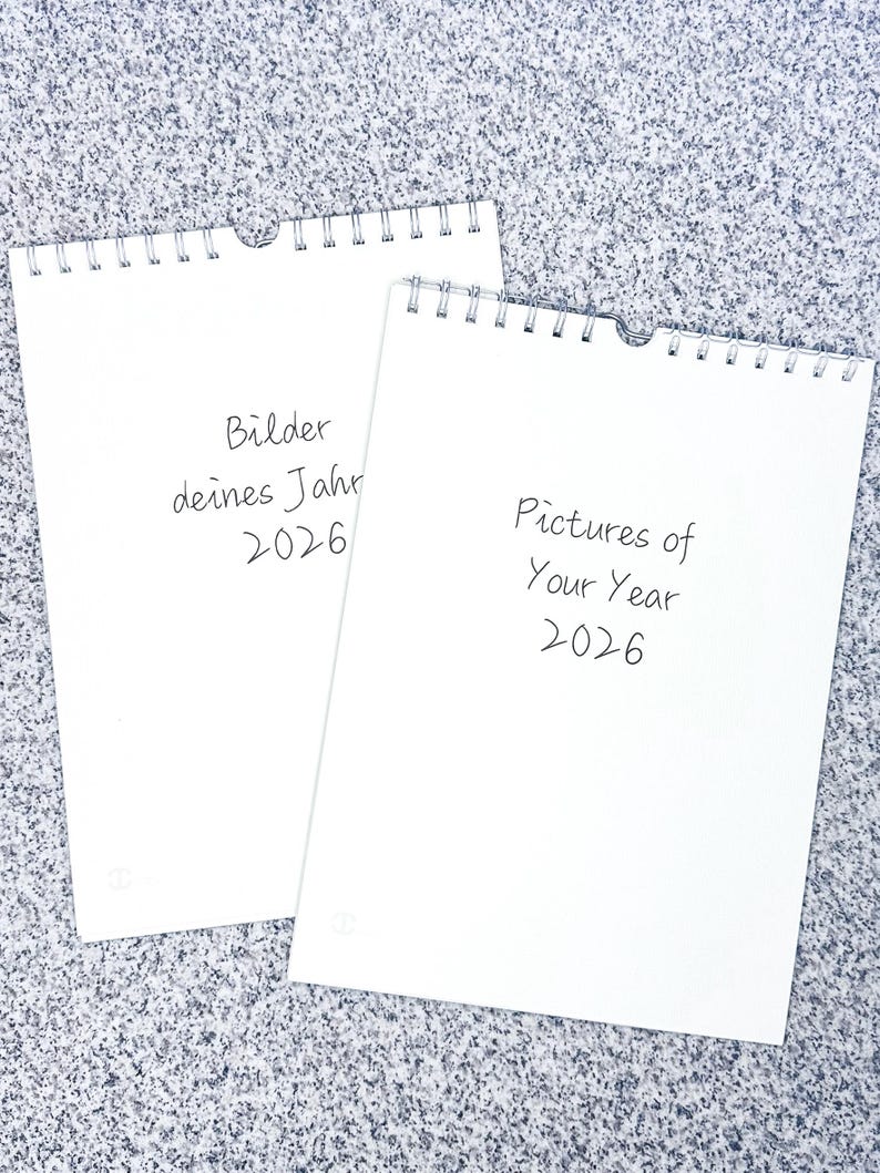 Op de afbeelding: Twee blanco witte kalenders met spiraalbinding. De bovenste kalender heeft de tekst "Bilder deines Jahr 2026" en de onderste kalender heeft de tekst "Pictures of Your Year 2026".