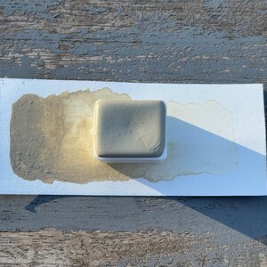May include: A square, gray watercolor paint pan sits on a white paper with a brown watercolor wash. The paint pan is in a white plastic holder. The paper is on a weathered, gray wooden surface.