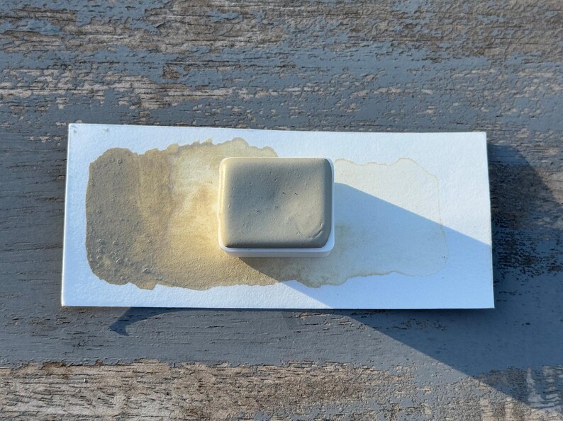 May include: A square, gray watercolor paint pan sits on a white paper with a brown watercolor wash. The paint pan is in a white plastic holder. The paper is on a weathered, gray wooden surface.