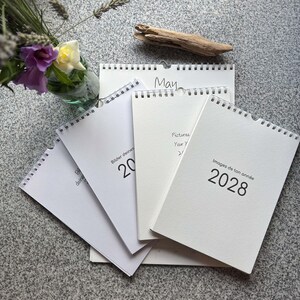 Mehrere Blankokalender gefächert: Maiseite und 2028-Cover auf Deutsch, Englisch und Französisch – mit Blumenstrauß und Treibholz auf Granit