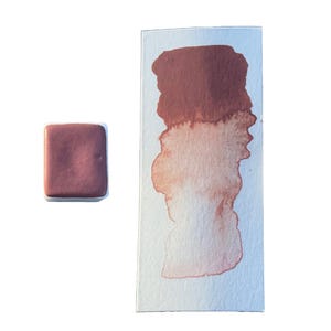 May include: A watercolor paint pan with a deep red-brown color. The paint is in a white pan with a square shape. A sample of the paint is shown on a white paper.
