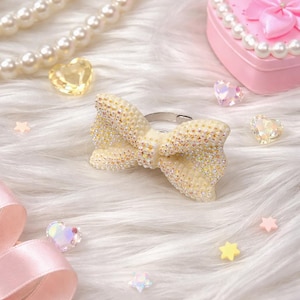 May include: A cream-colored bow ring adorned with sparkling crystals. The ring is set against a backdrop of white faux fur, with scattered heart-shaped and star-shaped embellishments. A pink heart-shaped box and a pearl necklace are also visible.