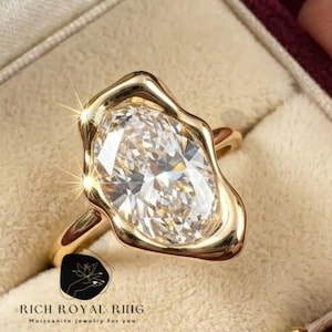 May include: A gold ring with a large, oval-shaped, clear gemstone. The gemstone is set in a unique, irregular bezel. The ring is displayed in a burgundy velvet ring box. The text "RICH ROYAL RING" is visible on the box.