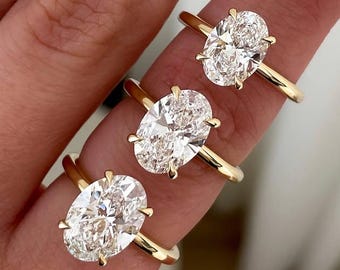 May include: Three gold rings with large, oval-cut diamonds are displayed on a finger. The rings have a simple, elegant design with a thin gold band. The diamonds sparkle brightly, reflecting light. The background is blurred, focusing attention on the jewelry.