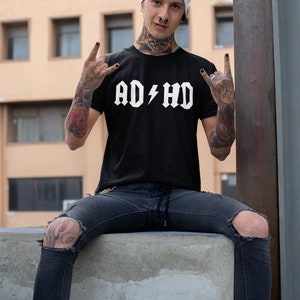 Peut inclure: Une personne portant un t-shirt noir avec le texte "AD/HD" en blanc, un jean d&eacute;chir&eacute; et un bonnet blanc. La personne a les cheveux verts et de nombreux tatouages.