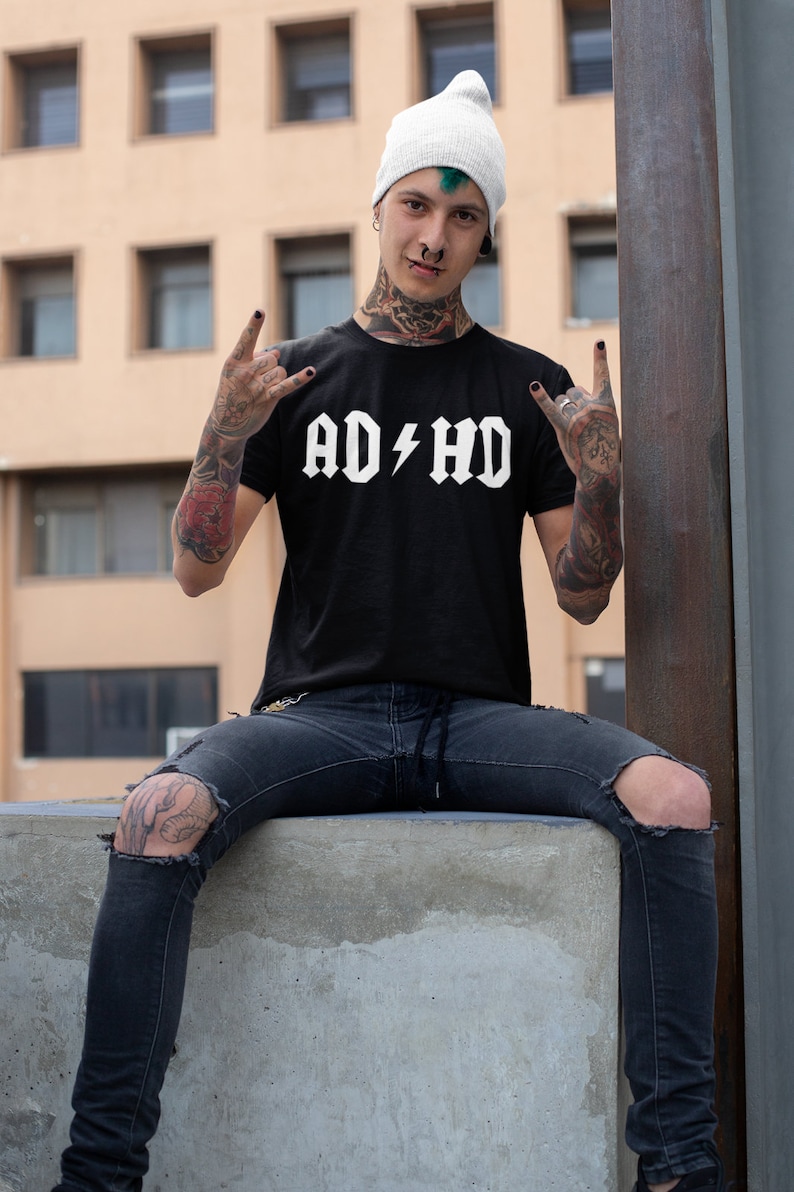 Peut inclure: Une personne portant un t-shirt noir avec le texte "AD/HD" en blanc, un jean d&eacute;chir&eacute; et un bonnet blanc. La personne a les cheveux verts et de nombreux tatouages.