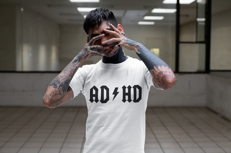 Peut inclure: Un t-shirt blanc avec un texte noir qui dit "AD/HD" avec un &eacute;clair entre les mots.