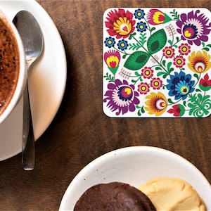 May include: A white square coaster featuring a colourful floral design. The design incorporates red, yellow, blue, green and purple flowers with white accents. The coaster is ideal for protecting surfaces from heat and spills.