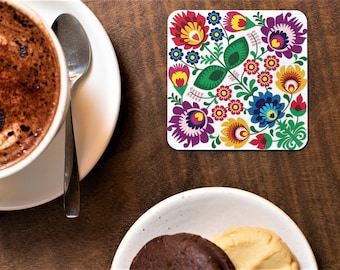 Puede incluir: Un posavasos cuadrado blanco con un diseño floral colorido. El diseño presenta flores rojas, amarillas, azules, verdes y moradas con detalles blancos. El posavasos es perfecto para proteger las superficies del calor y los derrames.