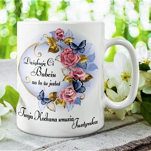 Puede incluir: Taza de cerámica blanca con un diseño azul y dorado con rosas y mariposas. La taza tiene el texto "Dziekuje Ci Babciu za to ze jestes Twoja Kochana wnuczka Justynka" impreso en ella.