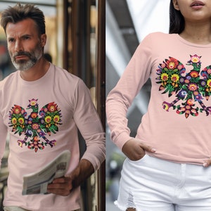 Può includere: Una maglietta rosa a maniche lunghe con un design floreale di un'aquila polacca. L'aquila è un simbolo tradizionale della Polonia.