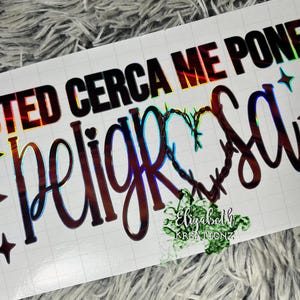 May include: A white sticker with the Spanish phrase "Usted Cerca Me Pone Peligrosa" in black and holographic lettering. The word "Peligrosa" is in a cursive font with a heart-shaped design. Decorative star shapes are also included.