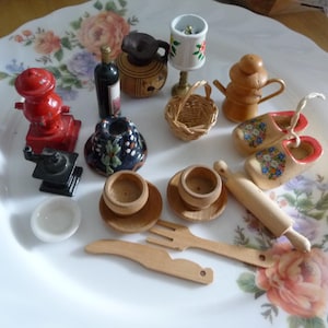 Op de afbeelding: Een verzameling miniatuur decoratieve items, waaronder een rode brandkraan, een zwarte koffiemolen, een kleine fles wijn en houten klompen met bloemmotieven. Andere items zijn een mand, een deegroller en een kleine lamp.