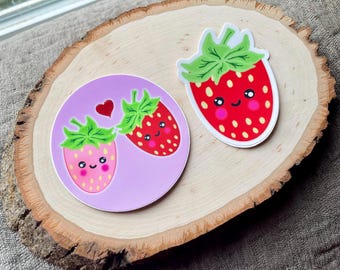 May include: Three strawberry stickers with cute faces. One sticker is a single red strawberry with white seeds and a green top. Another sticker features two strawberries on a pink background with a heart. The stickers are on a wooden slice.