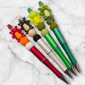 May include: A collection of five decorative pens with unique designs. Each pen features a different color scheme, including red, black, white, and green barrels. The pens are topped with various animal-themed embellishments, such as a red dragon and green alligators.