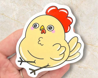 May include: A die-cut sticker featuring a cartoon chicken with a yellow body, red comb, and black and white eyes. The chicken has a cute, friendly expression and is outlined in black. The sticker is held in a hand.