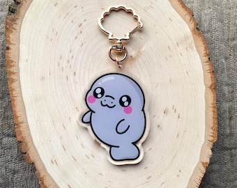 May include: A clear acrylic keychain featuring a cartoon manatee design. The manatee is light blue with pink cheeks and a smiling expression. The keychain has a gold-colored clasp and ring.