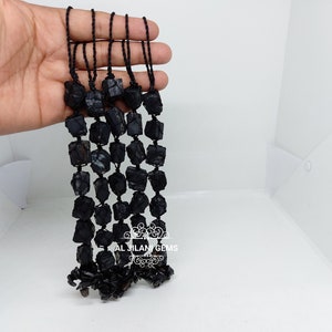 May include: Strands of black tourmaline gemstones. Each strand comprises raw, irregularly shaped black tourmaline stones threaded on black cord. The stones have a rough, natural texture and vary in size and shape. The background is white.