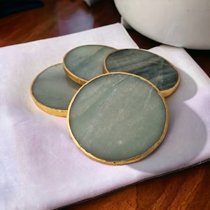 May include: Four circular coasters with a gold-coloured rim. The coasters have a light green and grey marbled appearance. They are arranged on a white cloth, with a blurred background.