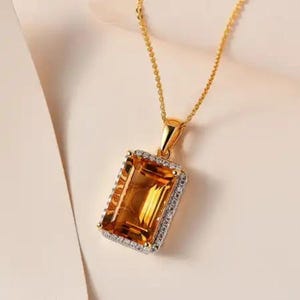 May include: A gold necklace with a rectangular, amber-colored pendant. The pendant is framed with small, clear stones. The chain and pendant setting are gold-toned, creating a luxurious look. The necklace is displayed against a soft, neutral background.