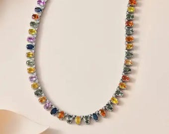 May include: A multi-colored gemstone necklace with oval-shaped stones in a variety of colors, including yellow, orange, blue, and purple. The necklace is set on a silver chain and is displayed against a light pink background.
