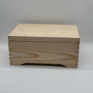 Puede incluir: Una caja de madera simple con una tapa con bisagras y una base ligeramente curvada. La caja está hecha de madera clara y tiene un acabado natural.