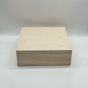 May include: A white wooden box with a hinged lid. The box is made of natural wood and has a light, unfinished look. The box is approximately 25 cm long, 20 cm wide, and 10 cm tall.