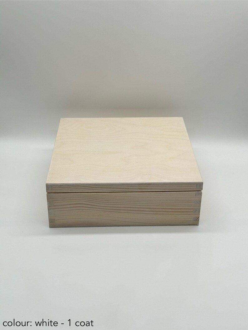 May include: A white wooden box with a hinged lid. The box is made of natural wood and has a light, unfinished look. The box is approximately 25 cm long, 20 cm wide, and 10 cm tall.