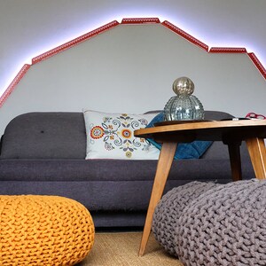 May include: A living room with a gray couch, two knitted poufs, one orange and one gray, a wooden coffee table, and a decorative glass orb. The wall behind the couch has a red LED light strip in the shape of a mountain range.