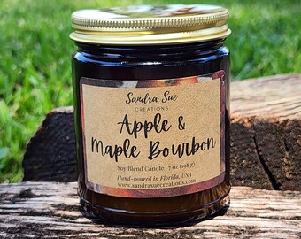 Puede incluir: Una vela de vidrio marrón en un frasco con tapa dorada. La etiqueta es marrón con texto blanco que dice "Sandra Sue Creations Apple & Maple Bourbon Soy Blend Candle | 7 oz (198 g) Hand-poured in Florida, USA www.sandrasuecreations.com".