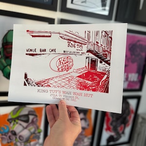 May include: A red and white print of King Tut's Wah Wah Hut, a music venue in Glasgow, Scotland. The print features the venue's name, address, and a stylised image of the building.