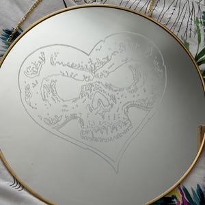 May include: Circular mirror with a gold frame and hanging chain. The mirror has an etched design of a skull within a heart shape. This decorative mirror is designed for wall mounting.