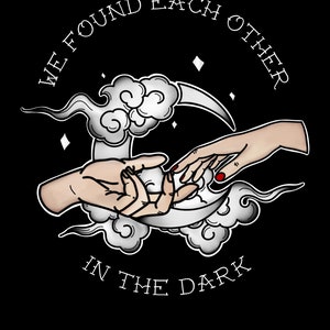 Può includere: Illustrazione in bianco e nero di due mani che si raggiungono, formando una forma di mezzaluna. Le mani sono circondate da nuvole e il testo "We found each other in the dark" è scritto in un cerchio attorno alle mani. La firma dell'artista e l'anno 2022 sono scritti sotto le mani.
