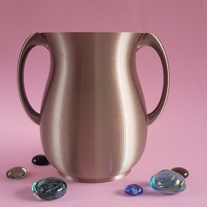 Puede incluir: Un jarrón de metal de color plateado con dos asas. El jarrón está sobre una superficie rosa con varias piedras pequeñas de vidrio de colores alrededor.