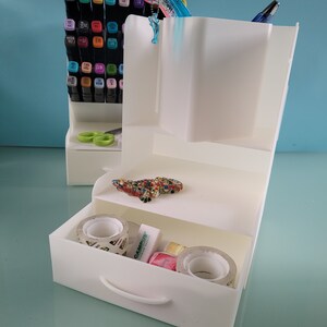 May include: White plastic desk organizer with a drawer, two shelves, and a pen holder. The organizer is filled with various office supplies, including pens, markers, tape, and a small toy lizard.