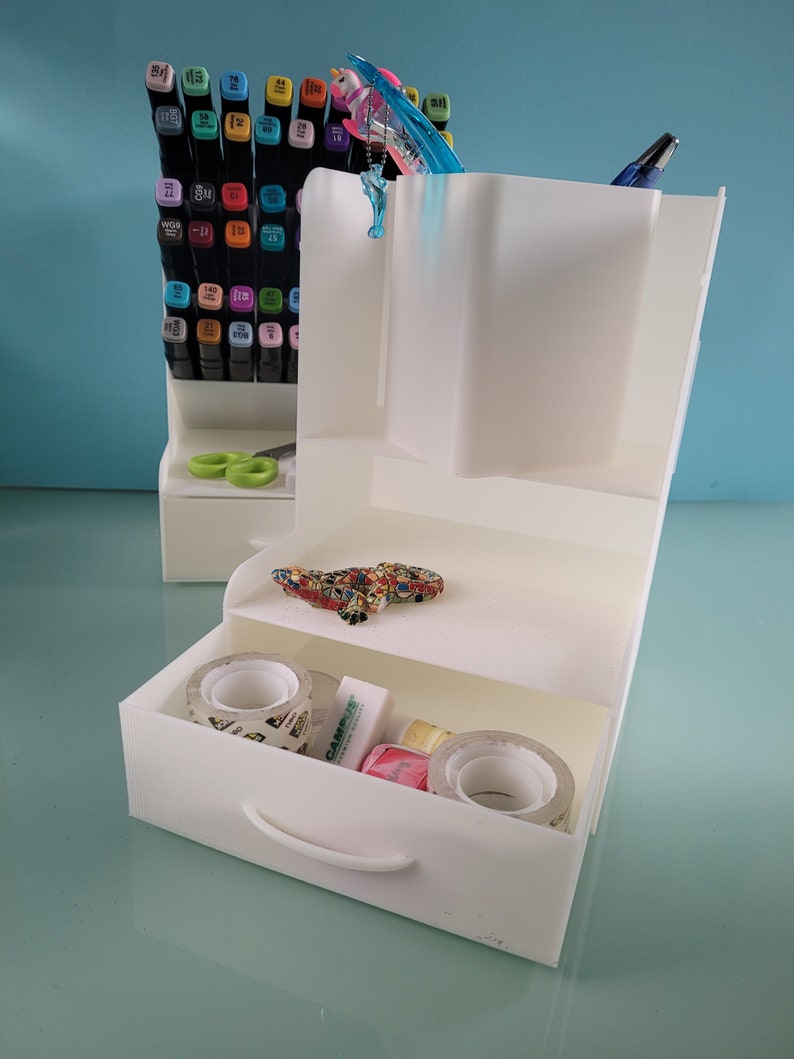 May include: White plastic desk organizer with a drawer, two shelves, and a pen holder. The organizer is filled with various office supplies, including pens, markers, tape, and a small toy lizard.
