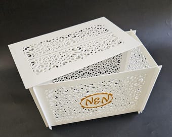 Op de afbeelding: Witte decoratieve doos met een opengewerkt ontwerp. De doos heeft een rechthoekige vorm met een gedeeltelijk open deksel. De doos heeft de initialen "N&N" in een gouden ovaal op de voorkant.