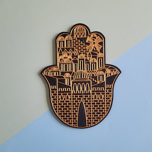 Puede incluir: Una decoración de pared de mano de Hamsa negra con un contorno dorado que representa un horizonte urbano con edificios y un molino de viento.