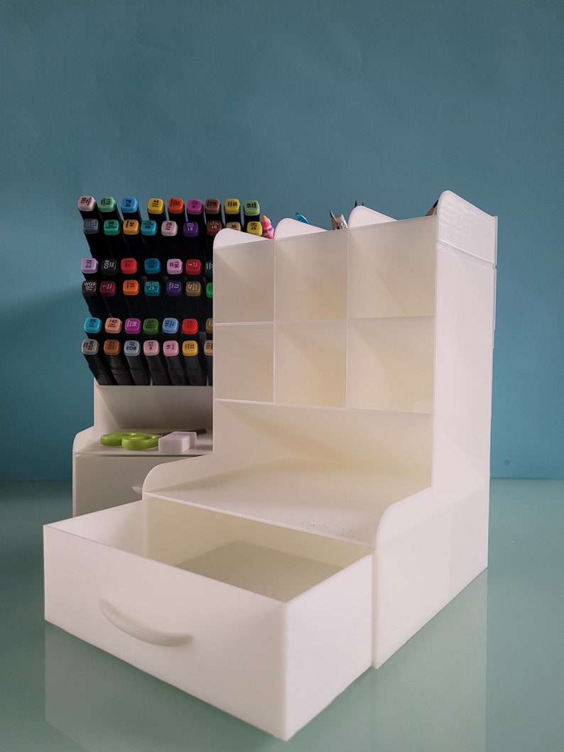 May include: White plastic desk organizer with a drawer and multiple compartments. The organizer is filled with colorful markers and pencils.