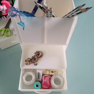 May include: White plastic desk organizer with two compartments for pens and pencils, a drawer with tape rolls, a pink eraser, a green eraser, and a small decorative lizard figurine.