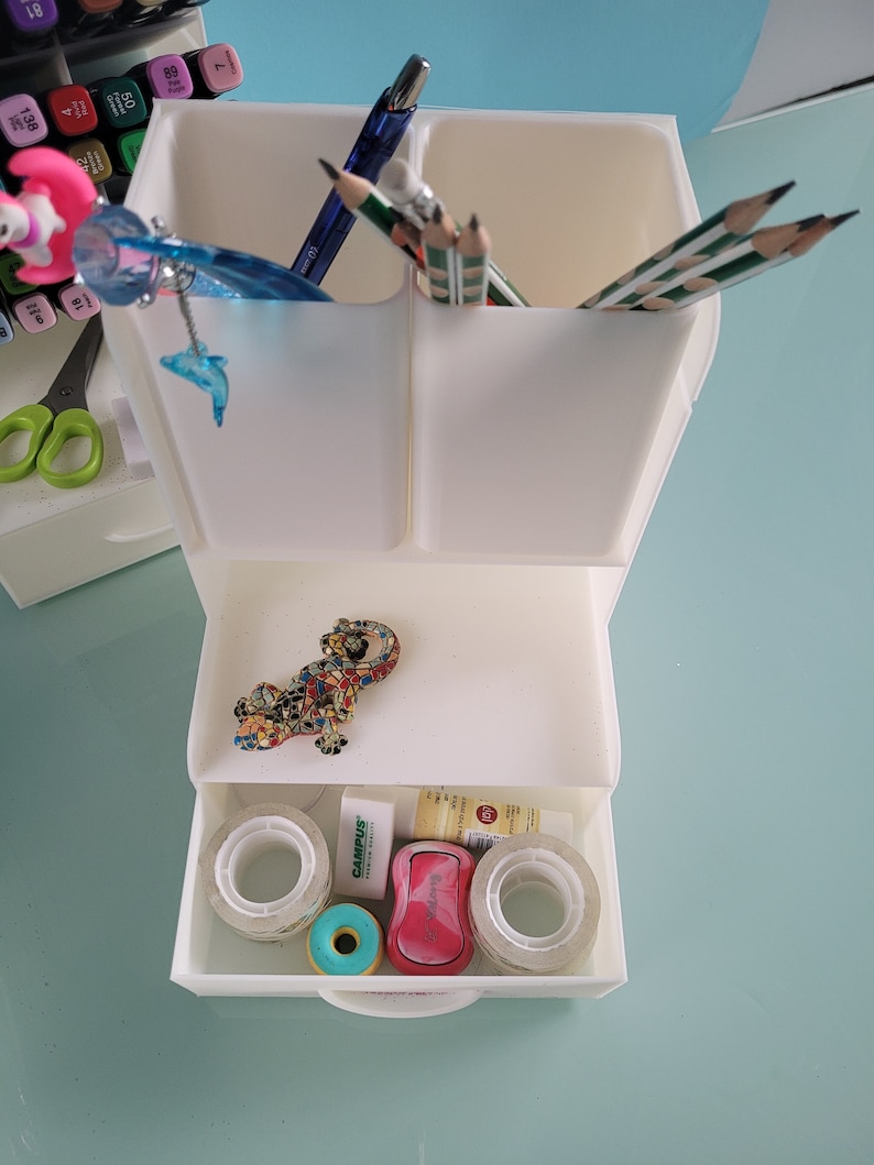 May include: White plastic desk organizer with two compartments for pens and pencils, a drawer with tape rolls, a pink eraser, a green eraser, and a small decorative lizard figurine.