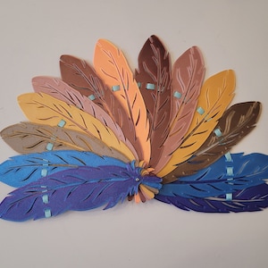 Puede incluir: Un abanico de plumas de papel de colores, dispuestas en semicírculo. Las plumas son en tonos de marrón, naranja, amarillo y azul. Las plumas tienen pequeños detalles azules a lo largo de los bordes.