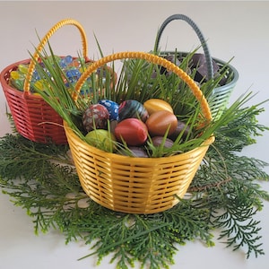 May include: Three yellow, red, and green wicker baskets filled with green grass and colorful Easter eggs. The baskets are sitting on a white surface with green pine branches.