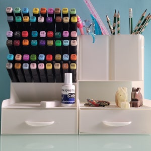 Puede incluir: Organizador de plástico blanco con dos cajones, un estante superior para bolígrafos y lápices, y un estante inferior para marcadores. El organizador está lleno de marcadores de colores, lápices y una botella de líquido blanco etiquetada como "Colorless Blending Solution".