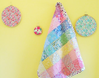May include: A colorful patchwork quilt hangs on a yellow wall, alongside three floral embroidery hoops. The quilt features squares of various floral patterns in pink, blue, green, and yellow. The embroidery hoops have floral fabric stretched across them, creating a decorative wall display.
