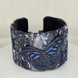 May include: A wide cuff bracelet with a textured, dark blue and silver exterior. A raised, detailed blue butterfly design is centered on the front. The interior is black.