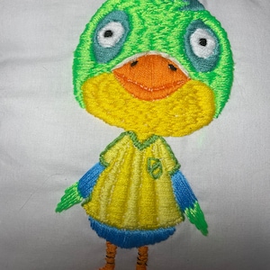 May include: Embroidered plush toy of a green and yellow duck wearing a yellow shirt with a blue collar and green trim. The duck has a large orange beak and blue eyes.
